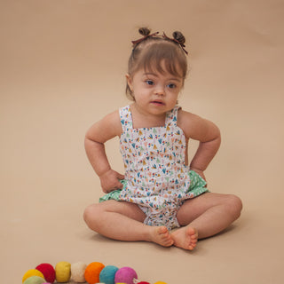 A toddler girl sits cross-legged on a beige studio floor wearing a sleeveless white romper with a colorful bird print and green ruffled shorts. She has brown hair in two small buns with bows.